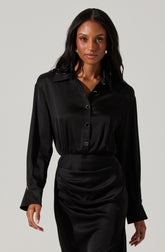 A woman is wearing a black satin button-up blouse with long sleeves and a collar. The blouse has a shiny texture and is slightly loose-fitting. She is also wearing a matching black satin skirt that is fitted at the waist with gathered detailing, creating a draped effect. The outfit appears elegant and sophisticated.